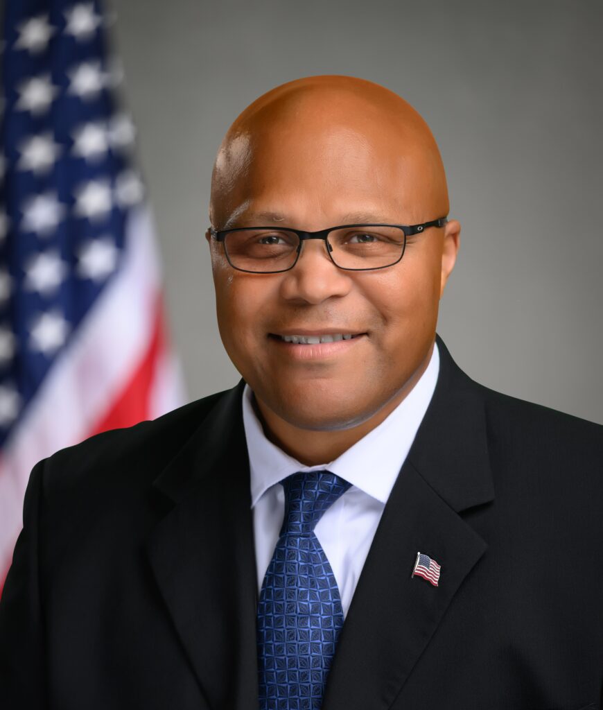 Photo of a man in a dark suit with a white button down and blue tie. There is a dark grey background and an American flag behind him.