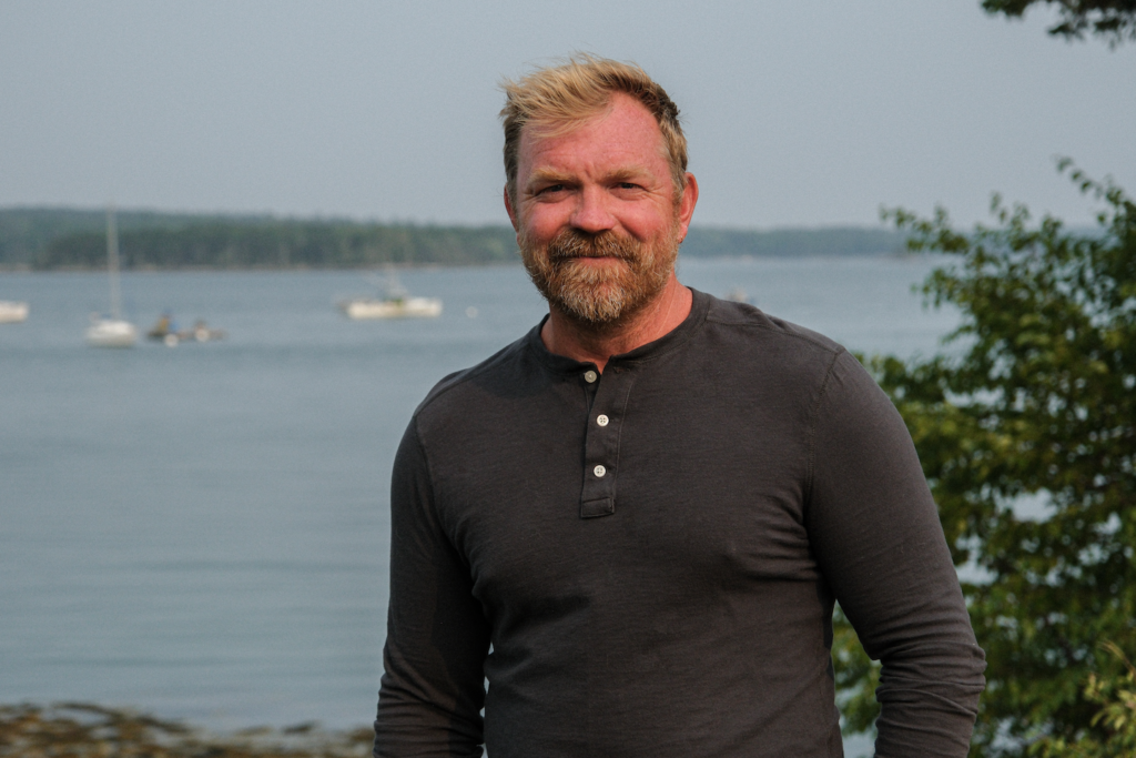 photo of a man in a dark henley shirt smiling with a body of water and trees behind him