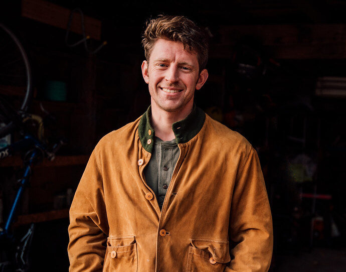 Photo of a man smiling with his hands in his front jean pockets, wearing a brown jacket