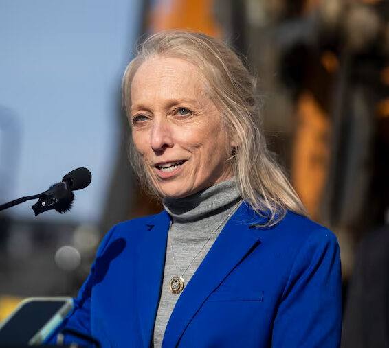 Photo of a woman in a blue blazer and grey turtleneck speaking into a microphone